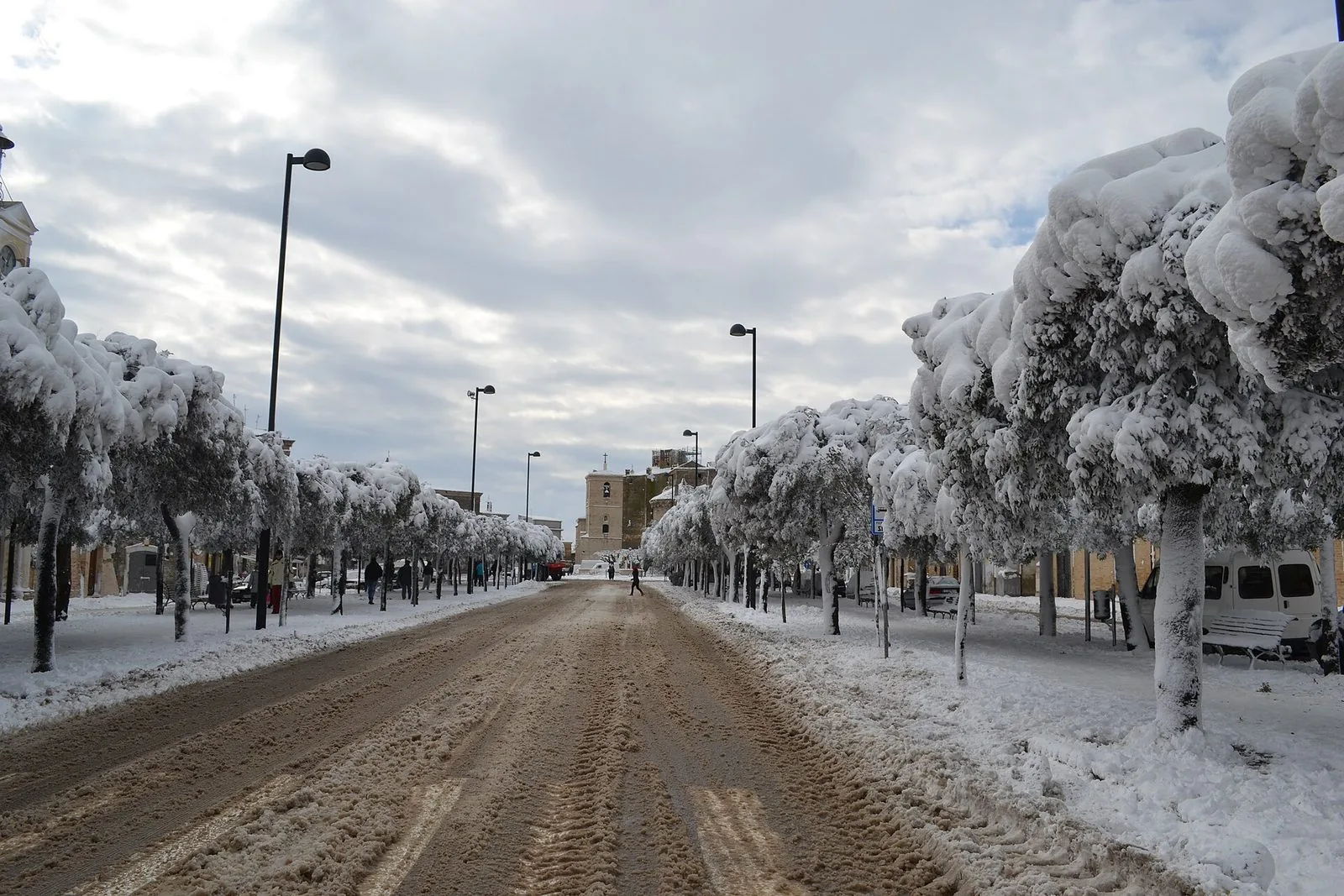 Il viale alberato di Serracapriola coperto di neve, con il campanile e la mole del castello in fondo alla strada sotto un cielo nuvoloso