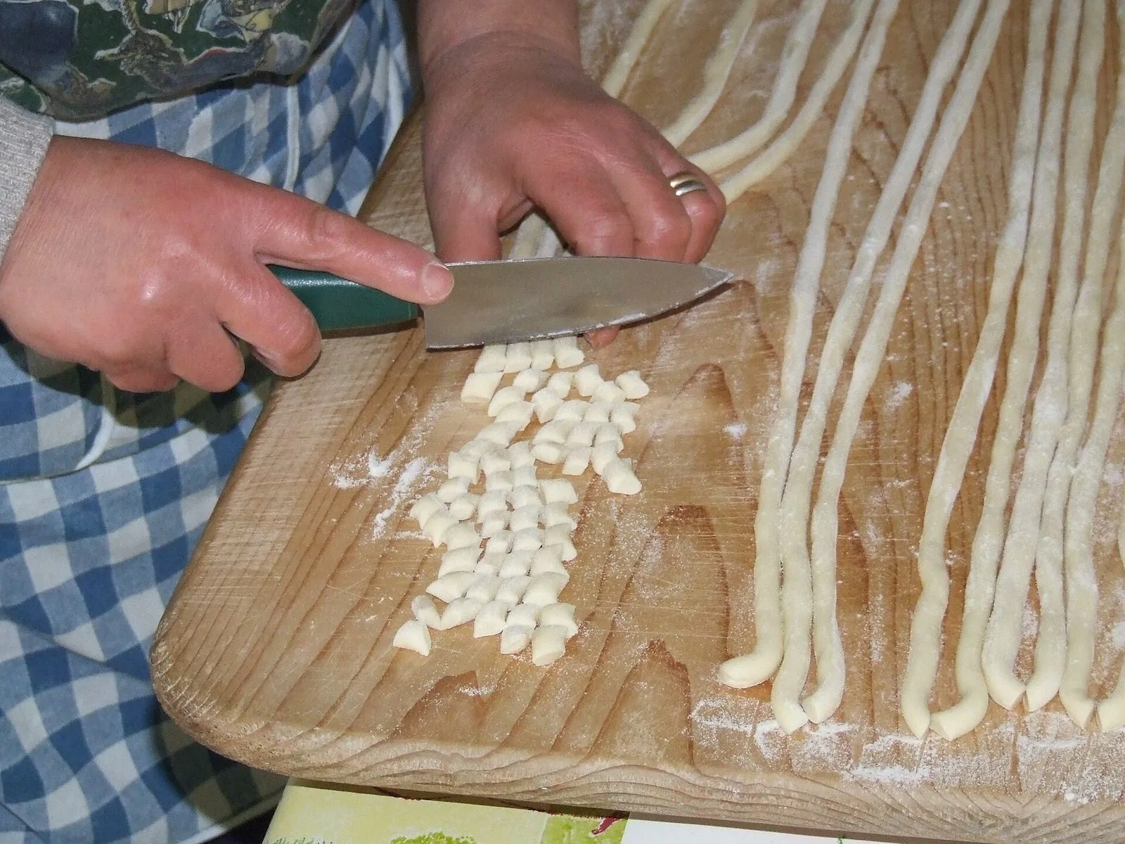 Due mani tagliano a tocchetti con un coltello dei cordoni di pasta fresca su una spianatoia di legno infarinata, accanto a un grembiule a quadretti bianchi e blu