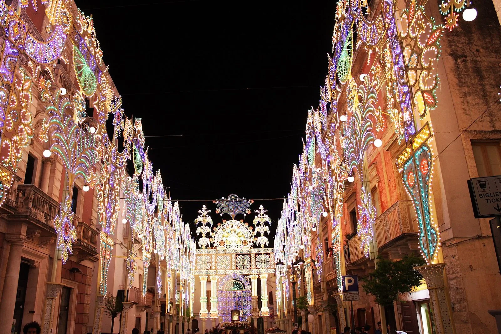 Una via di un centro storico pugliese di notte, interamente coperta da luminarie colorate per una festa patronale, con una grande galleria di luci sul fondo