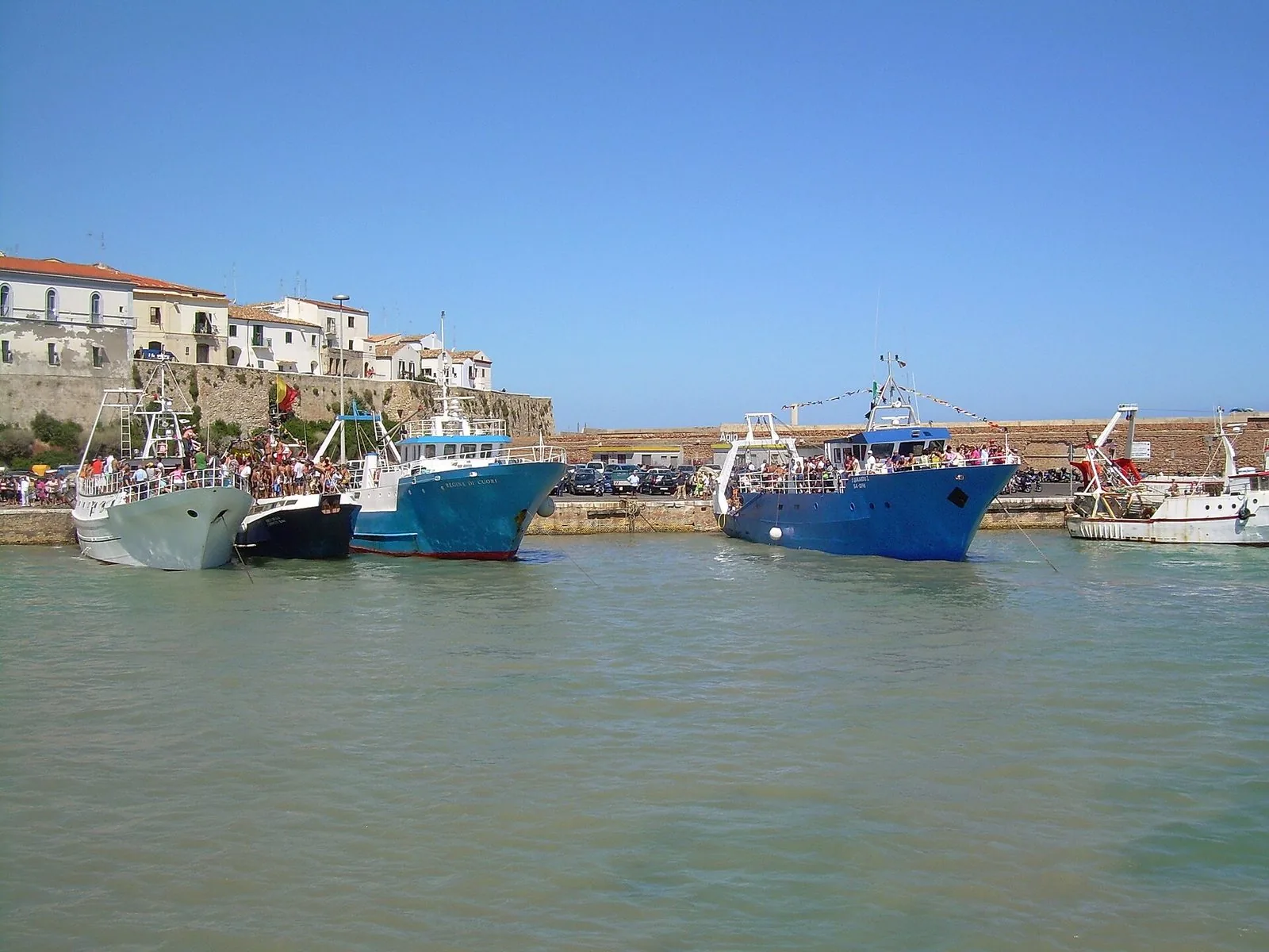 Pescherecci blu e bianchi ormeggiati nel porto di Termoli, con persone a bordo e sulla banchina, sotto le mura e le case bianche del borgo antico
