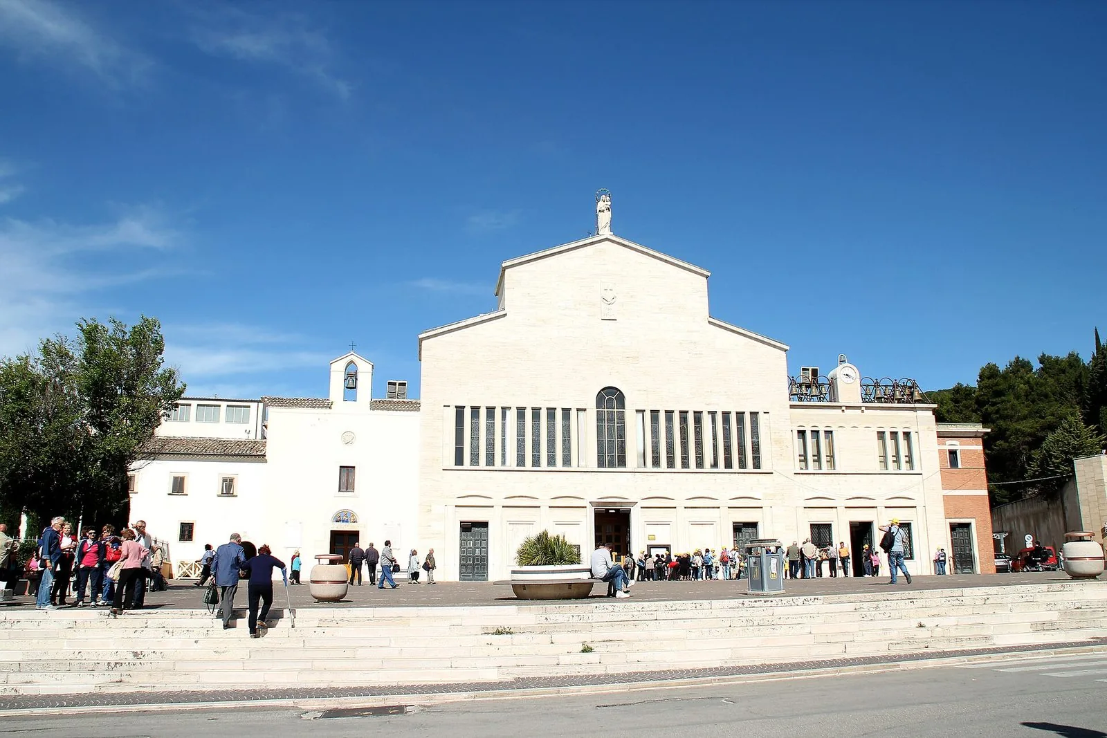 La facciata bianca della chiesa di Santa Maria delle Grazie accanto all'antico convento cappuccino di San Giovanni Rotondo, con pellegrini sul sagrato sotto un cielo azzurro