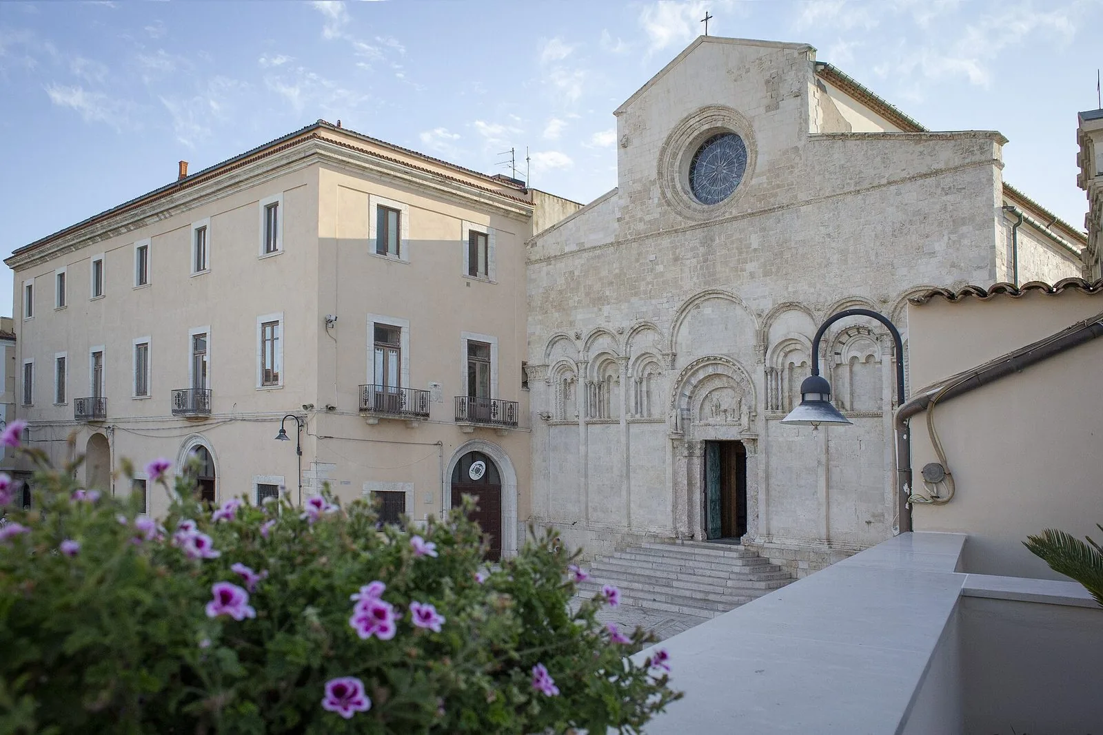La facciata romanica in pietra chiara della Cattedrale di Termoli con il rosone e gli archi ciechi, accanto a un palazzo color sabbia e fiori di geranio in primo piano